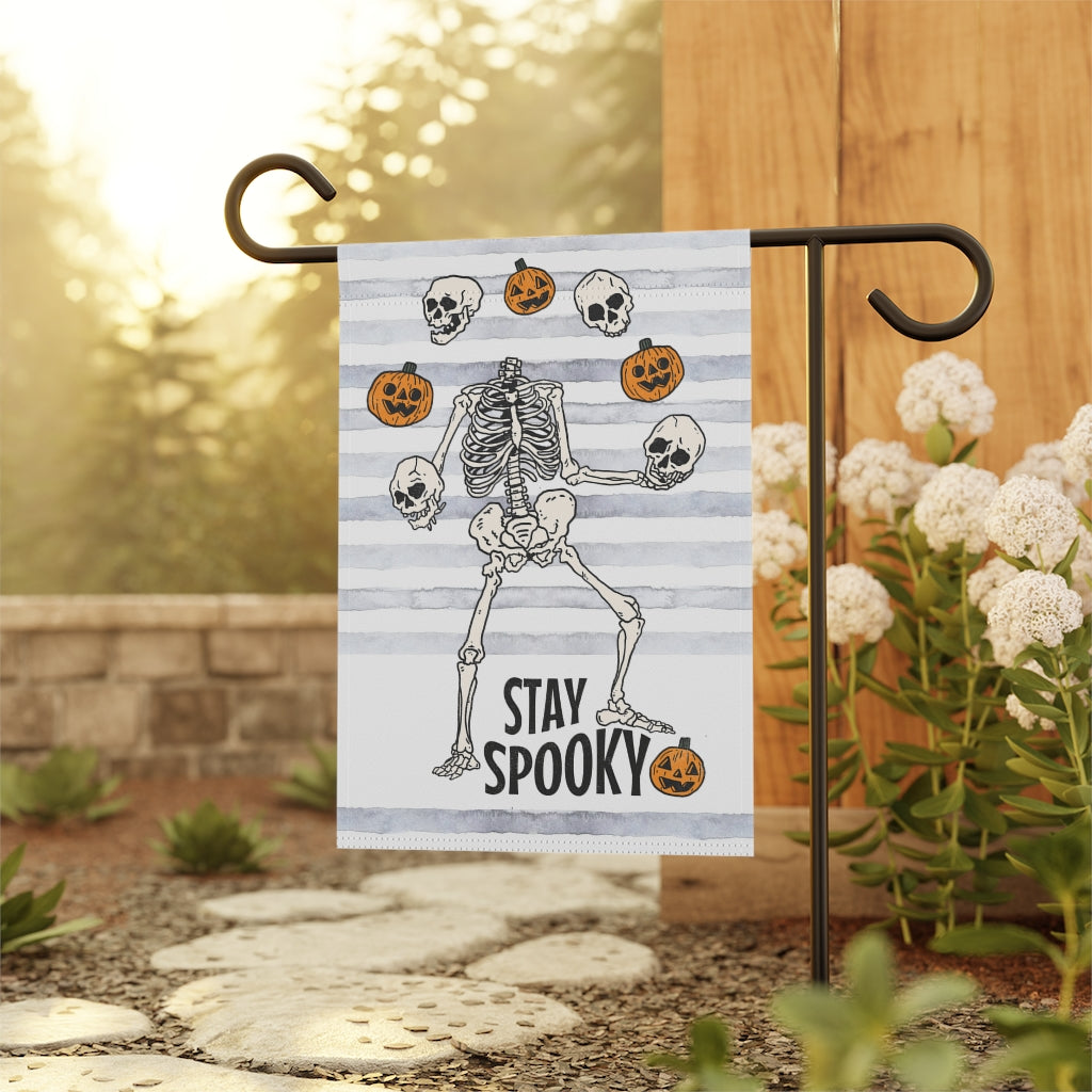 garden flag with dancing skeletons juggling pumpkins. The words on the bottom of the flag say stay spooky.