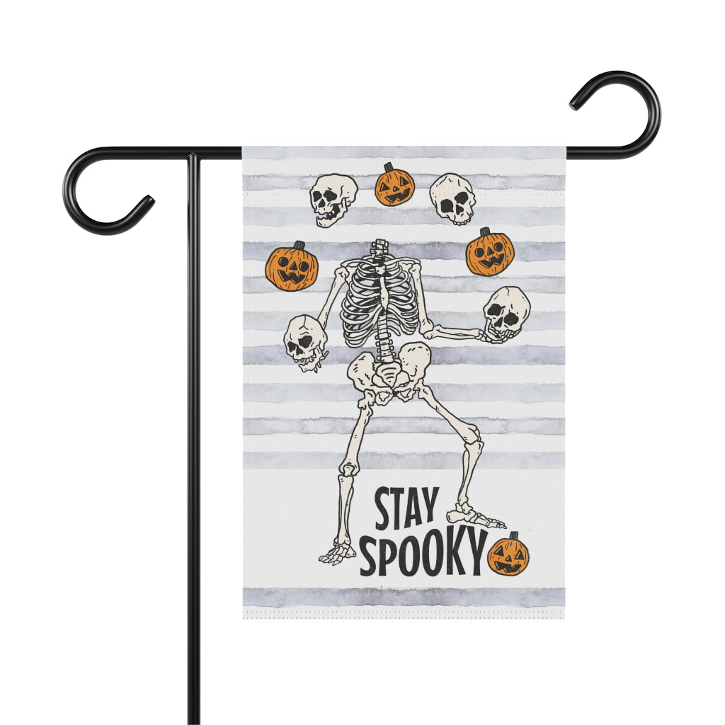garden flag with dancing skeletons juggling pumpkins. The words on the bottom of the flag say stay spooky.