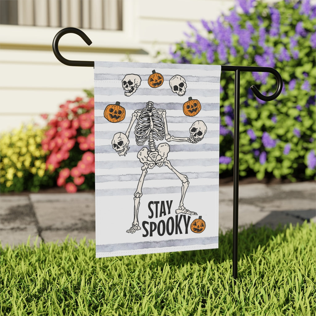 garden flag with dancing skeletons juggling pumpkins. The words on the bottom of the flag say stay spooky.