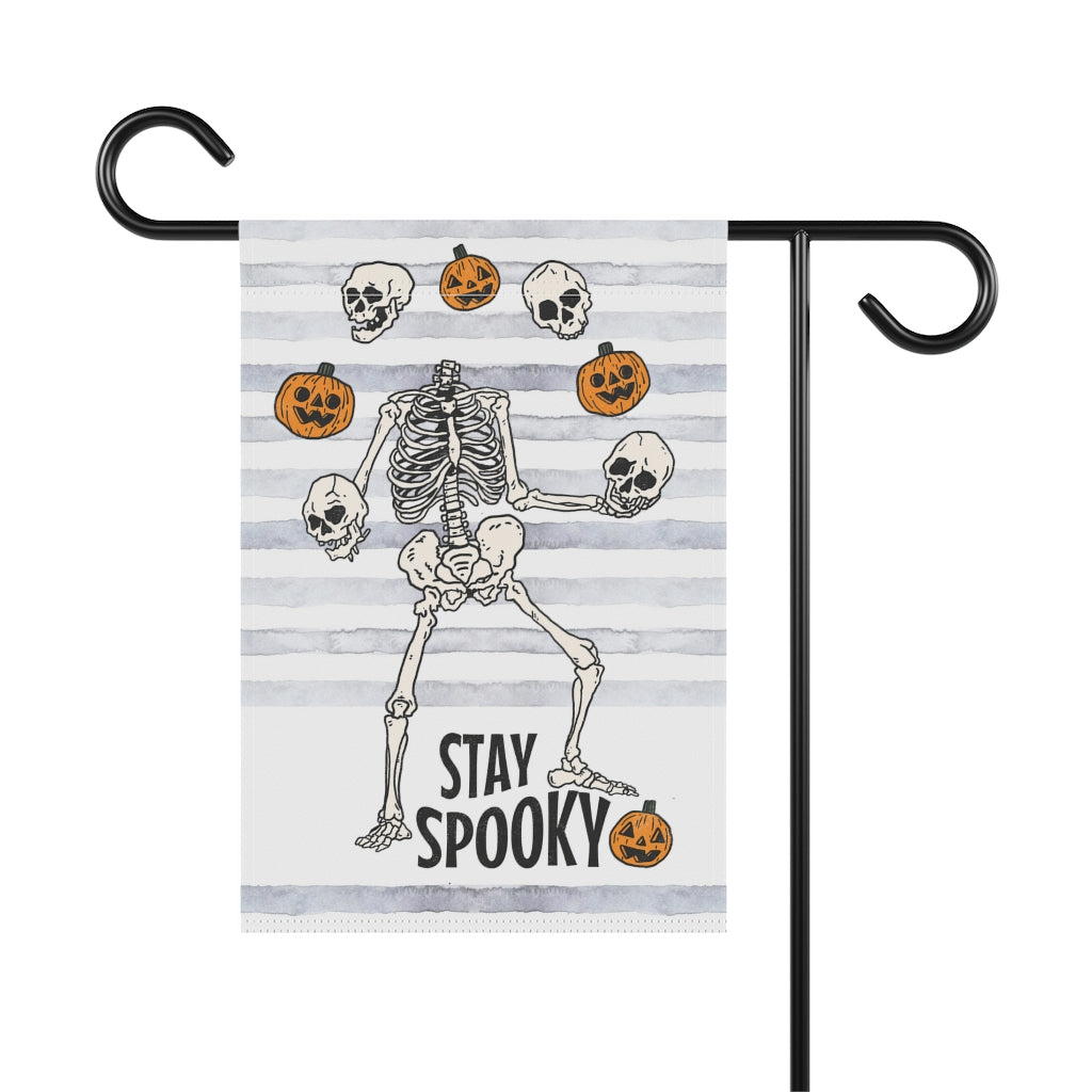 garden flag with dancing skeletons juggling pumpkins. The words on the bottom of the flag say stay spooky.